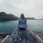 woman wearing gray long sleeved shirt and black black bottoms outfit sitting on gray wooden picnic table facing towards calm body of water at daytime